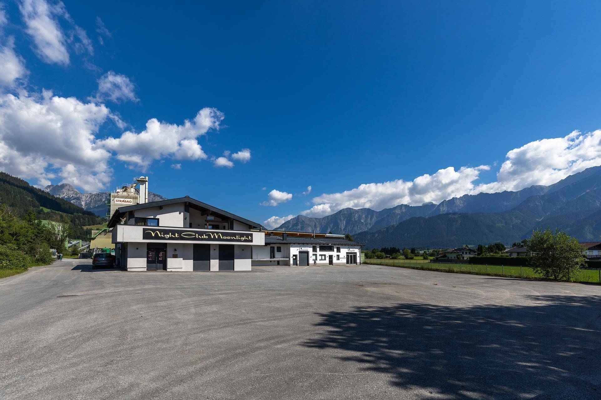 Gewerbegebäude "Night Club Moonlight" mit großem Parkplatz und beeindruckender Bergkulisse unter blauem Himmel.