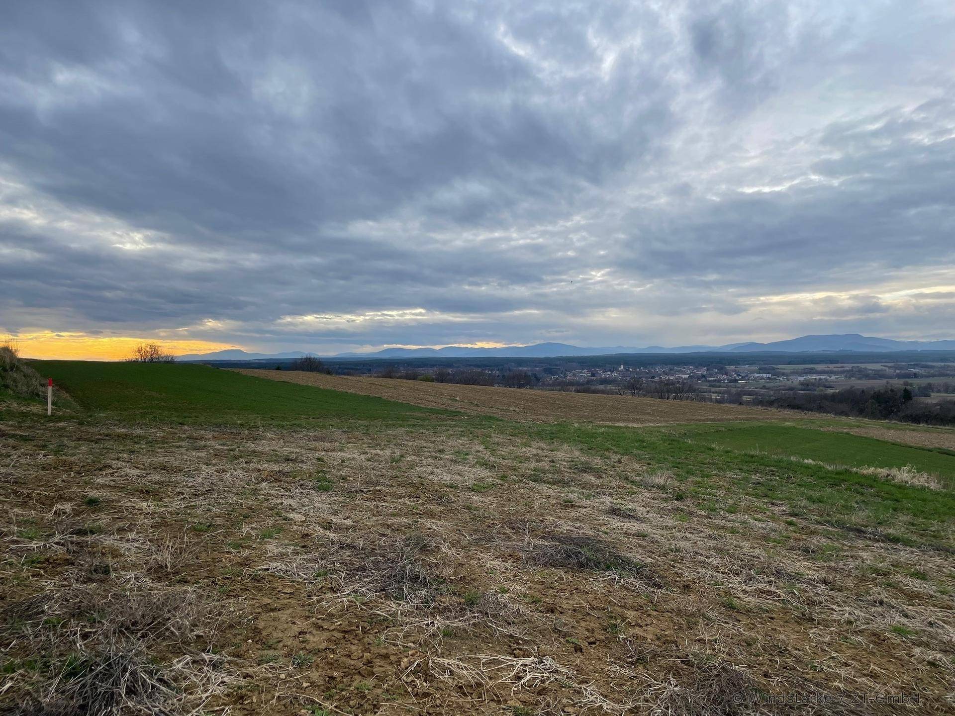 Weitläufige Landschaft mit Feldern und einem Dorf in der Ferne unter bewölktem Himmel.