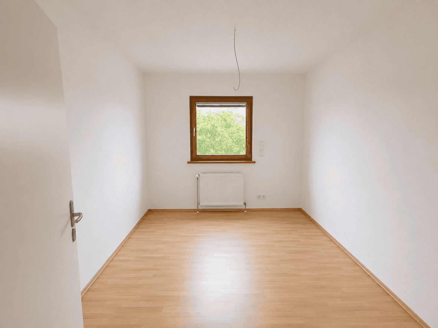 Helles Zimmer mit Laminatboden und einem großen Fenster mit Blick ins Grüne.
