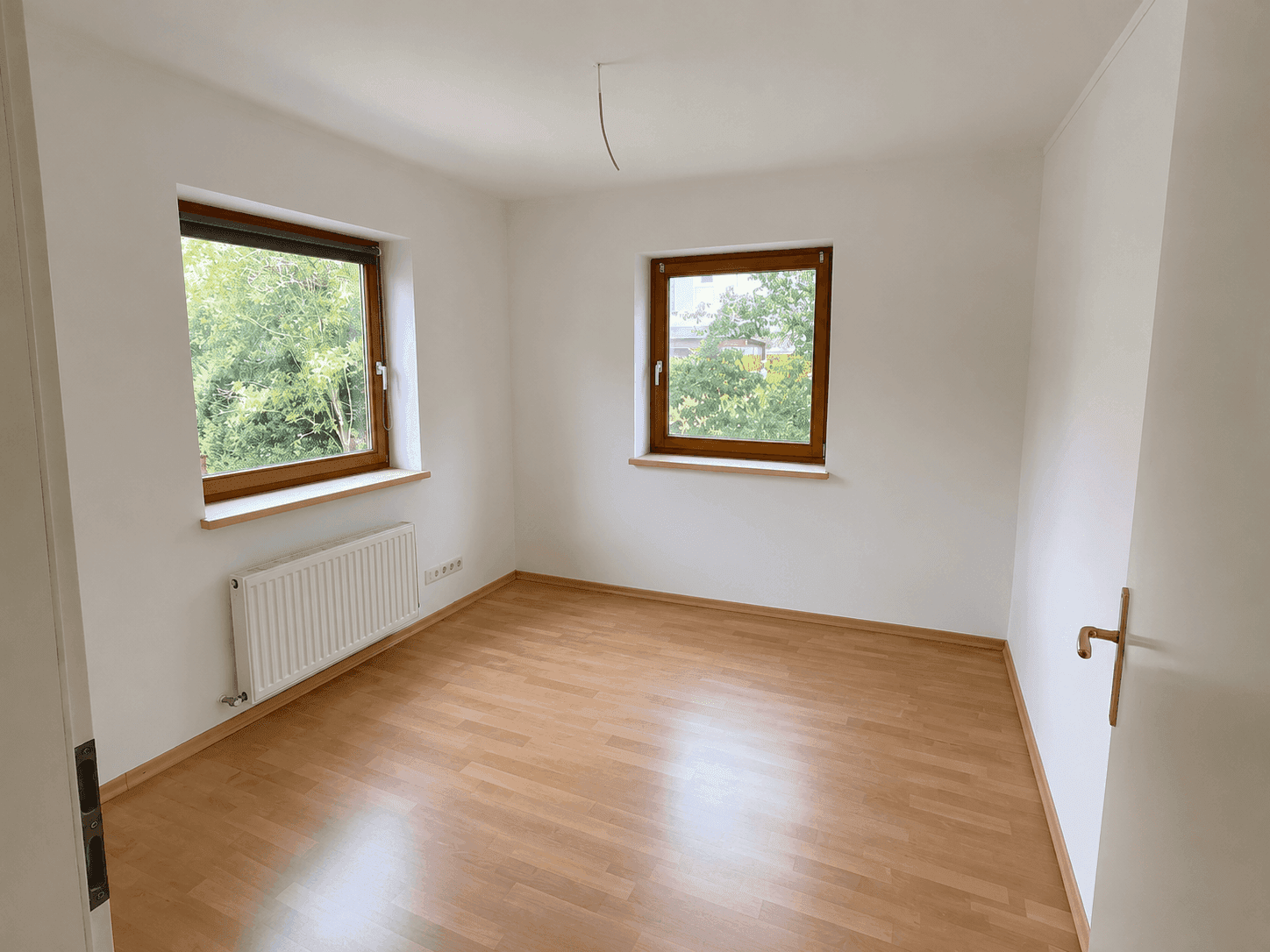 Helles Schlafzimmer mit Laminatboden und zwei Fenstern mit Blick ins Grüne.