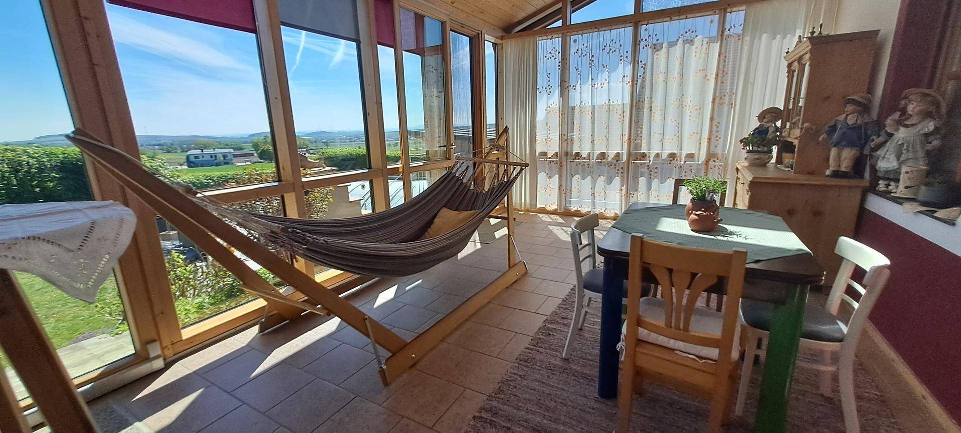 Heller Wintergarten mit Hängematte, Esstisch und weitem Blick über die Landschaft.