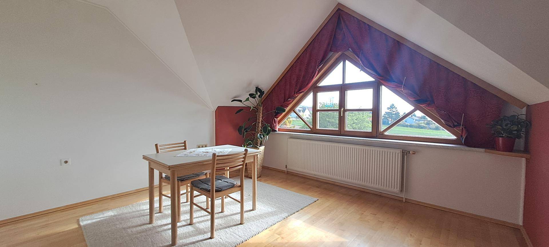 Helles Zimmer im Dachgeschoss mit Holzboden, schrägen Wänden und großem Giebelfenster.