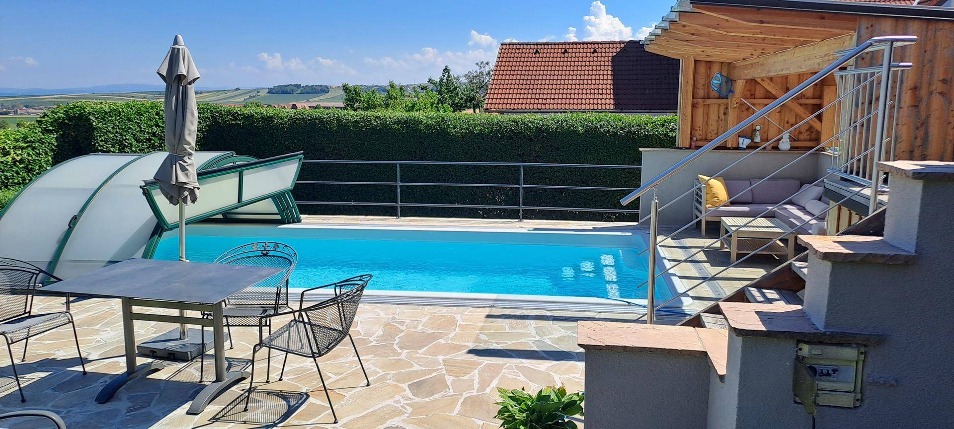 Entspannender Poolbereich mit Terrasse, Sonnenschirm und Zugang zum Haus über eine Treppe.