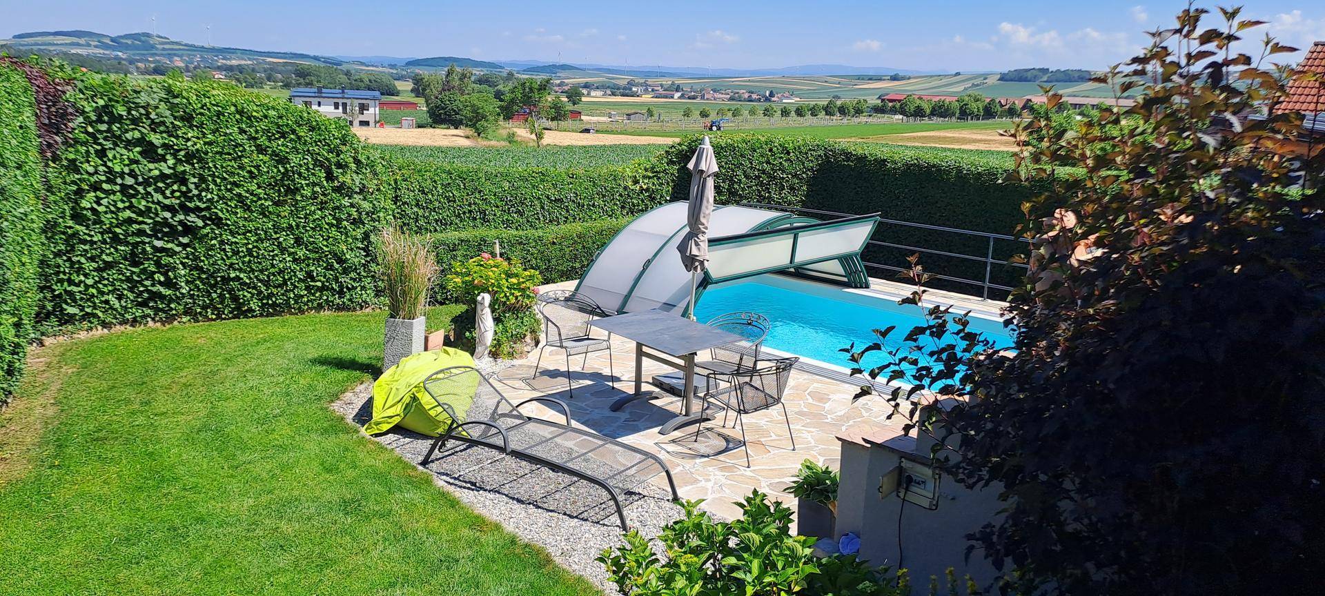 Sonniger Poolbereich im Garten mit Liegestühlen, Esstisch und weitem Landschaftsblick.