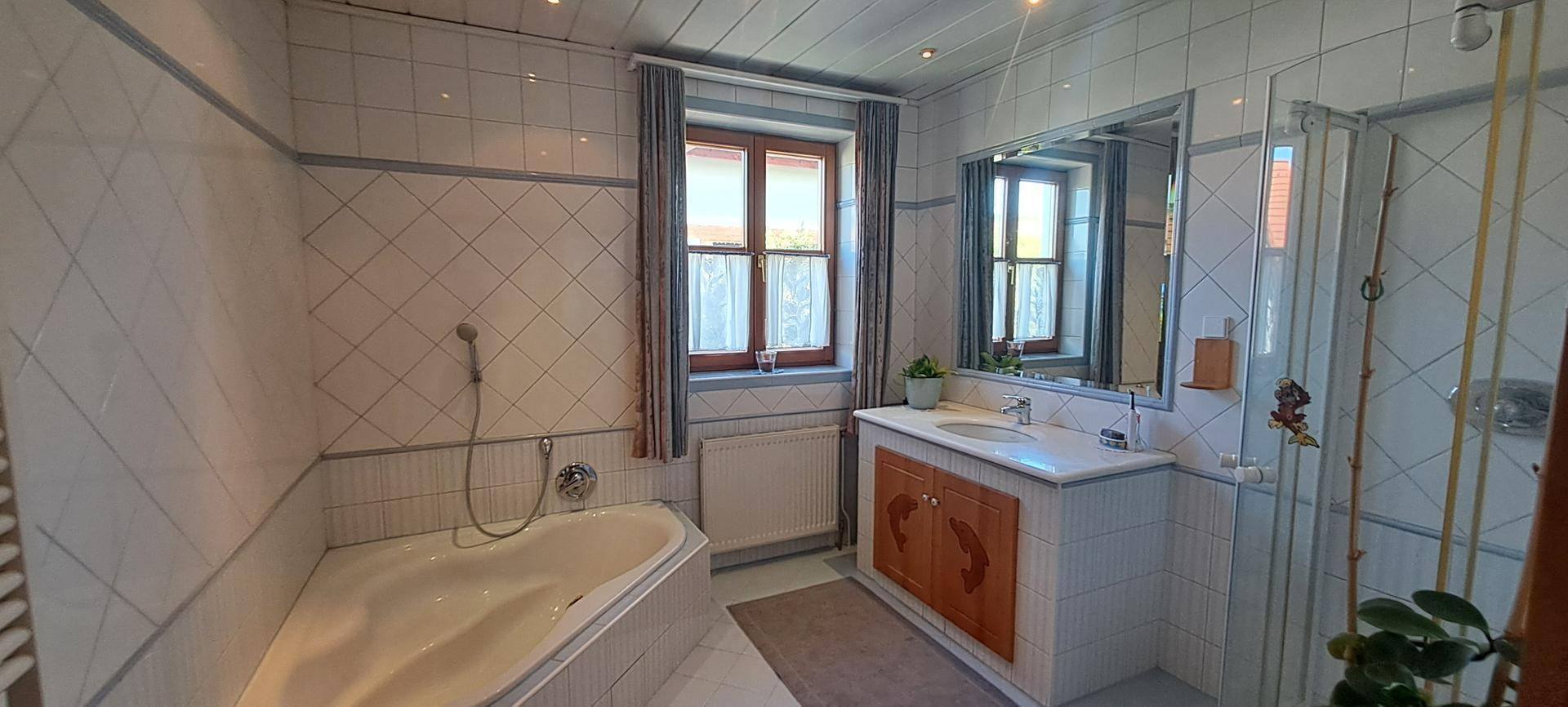Helles Badezimmer mit Badewanne, Dusche und Fenster, ausgestattet mit weißen Fliesen.