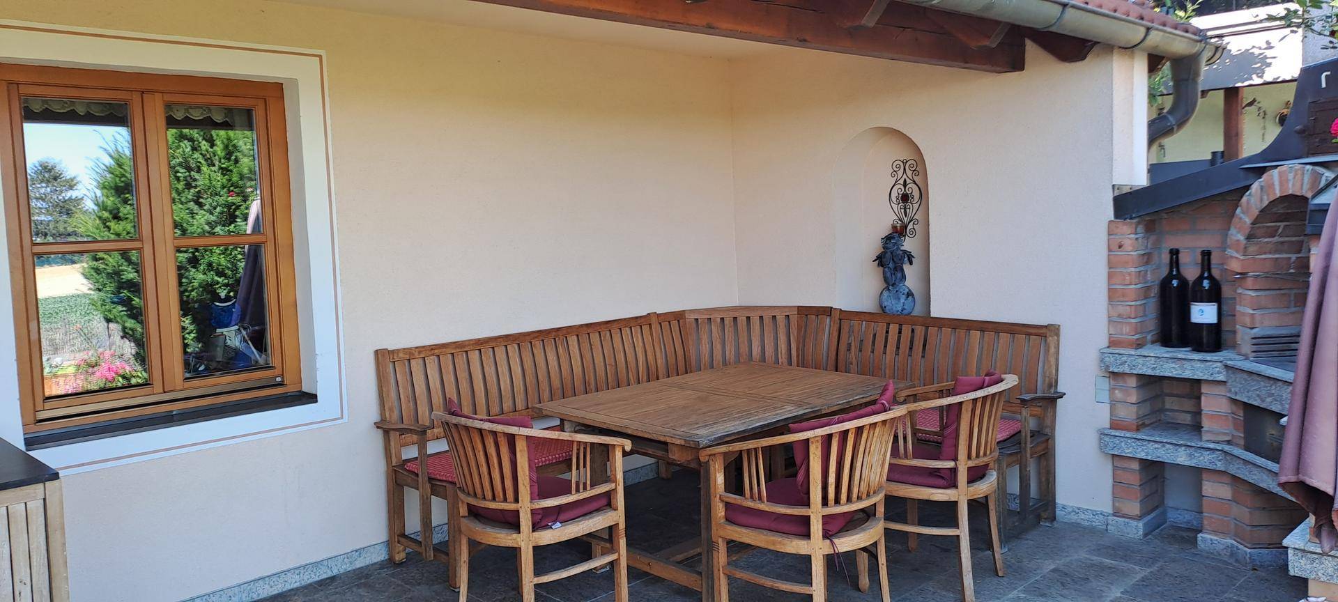 Überdachte Terrasse mit Holzbänken, Esstisch und gemauertem Grillbereich.