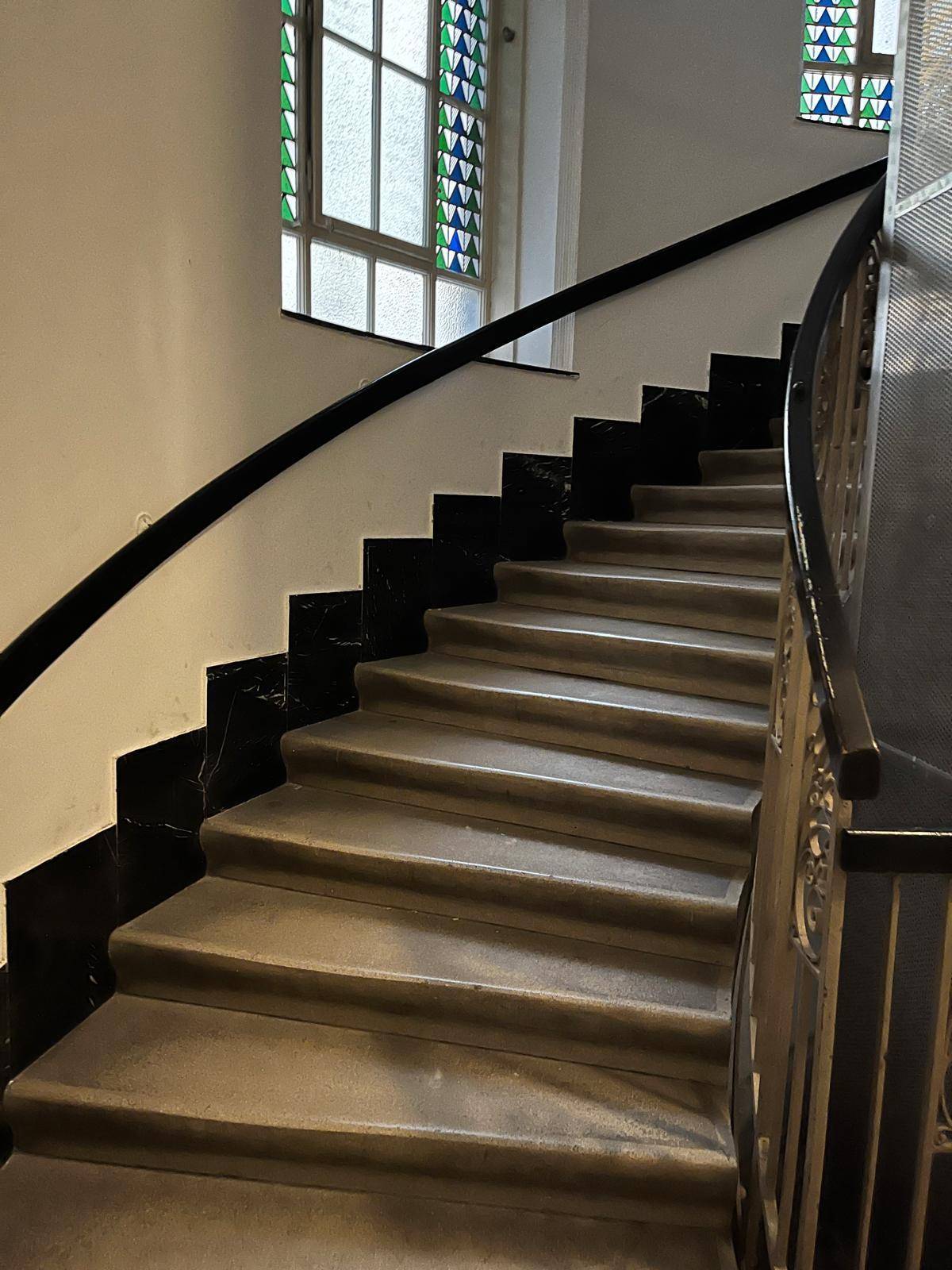 Geschwungene Treppe mit farbigen Glasfenstern und dunklem Handlauf in einem Altbau.