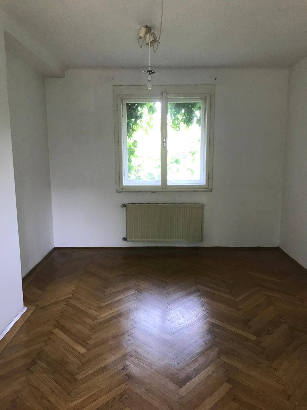 Leeres Zimmer mit Fischgrätparkettboden, einem Fenster und einem Heizkörper.