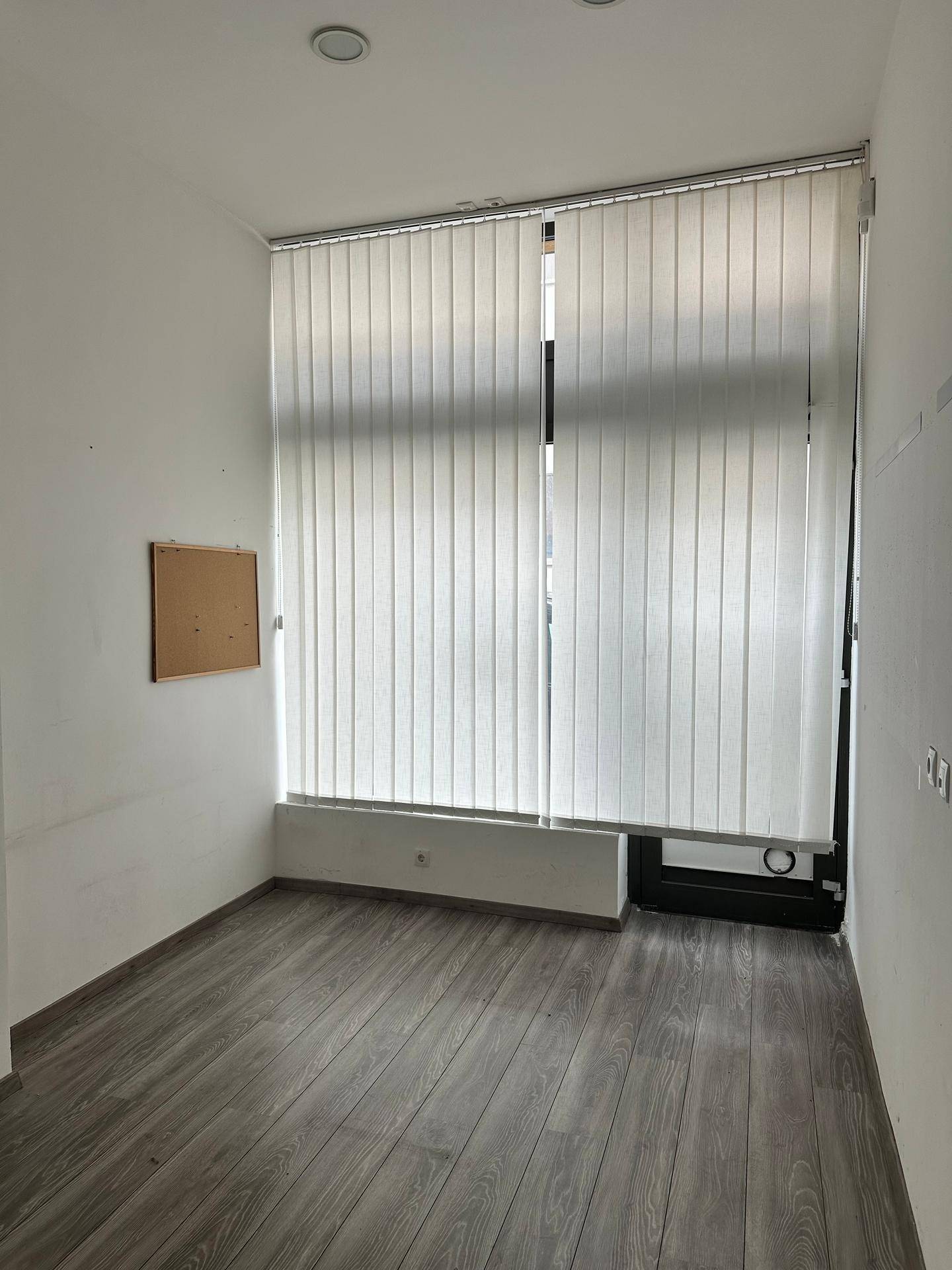 Leerer Büroraum mit großen Fenstern, vertikalen Jalousien und modernem Laminatboden.