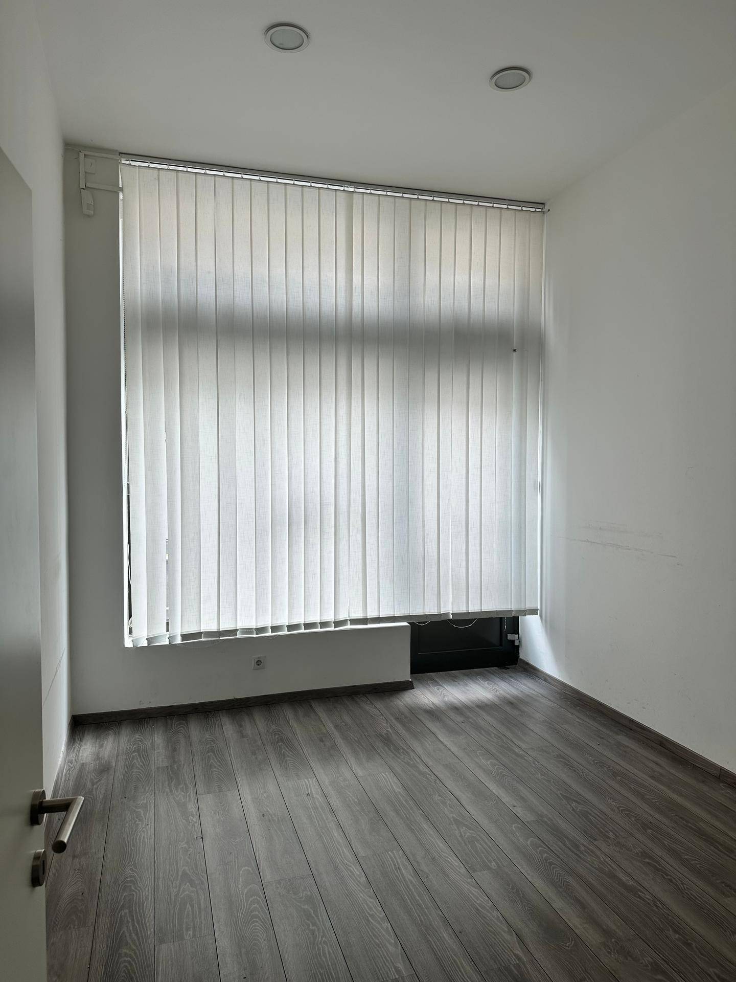Leerer Büroraum mit großen Fenstern, vertikalen Jalousien und modernem Laminatboden.