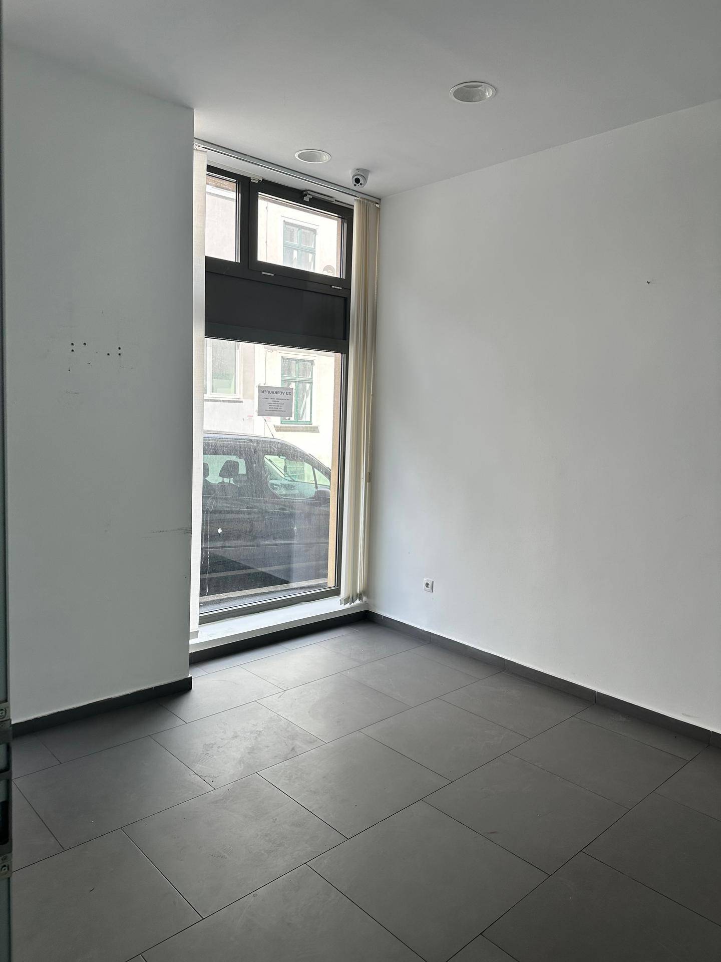 Leerer Büroraum mit großem Fenster zur Straße und dunklen Bodenfliesen, inklusive Überwachungskamera.