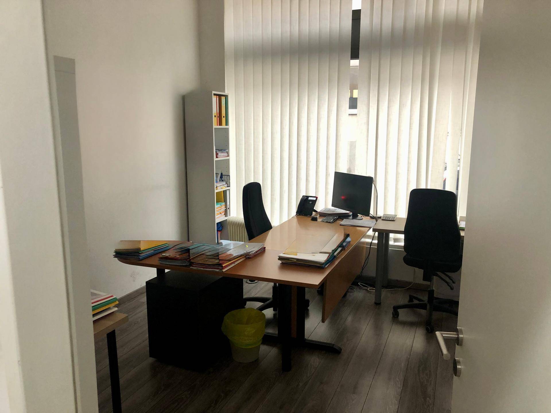 Helles Büro mit Schreibtischen, Bürostühlen und vertikalen Jalousien vor dem großen Fenster.