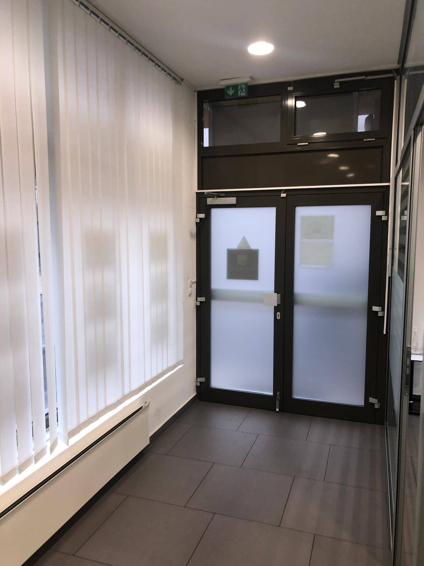 Eingangsbereich mit dunklen Fliesen, vertikalen Jalousien und einer modernen Glastür mit Milchglas.