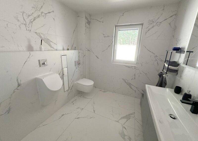 Modernes Badezimmer mit Marmoroptik-Fliesen, Urinal, wandhängendem WC und Fenster für natürliches Licht.