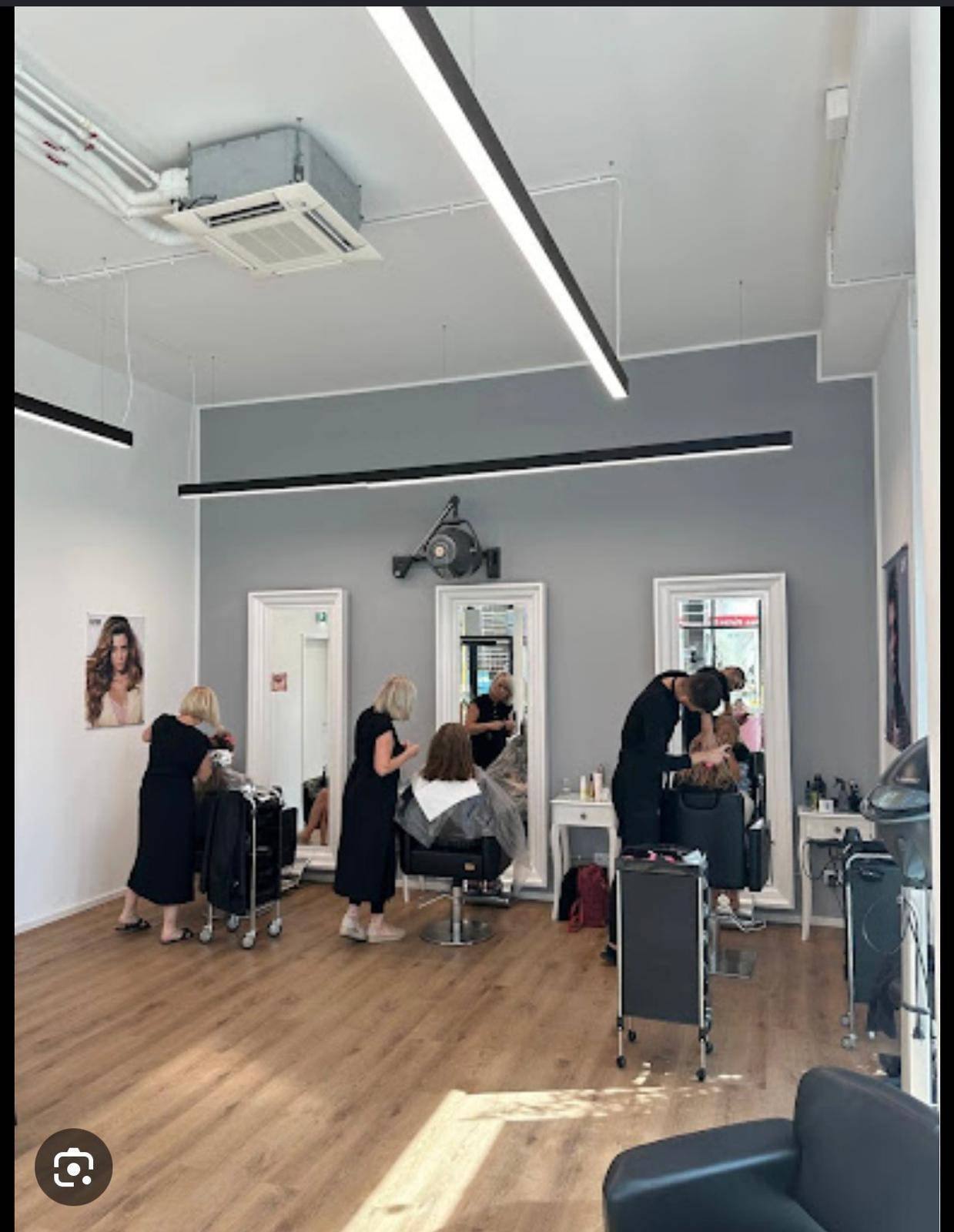 Innenansicht eines Friseursalons mit mehreren Bedienplätzen, grauen Wänden, Holzboden und sichtbarer Klimaanlage.