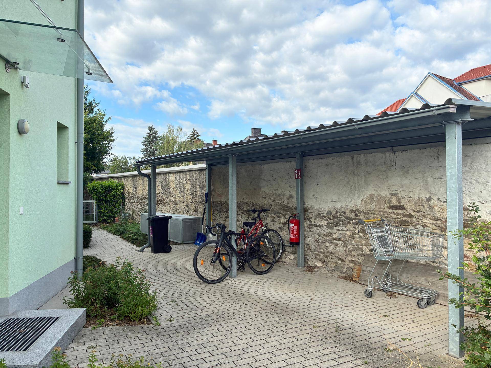 Überdachter Stellplatz mit Fahrrädern und Mülltonnen neben einer alten Steinmauer.