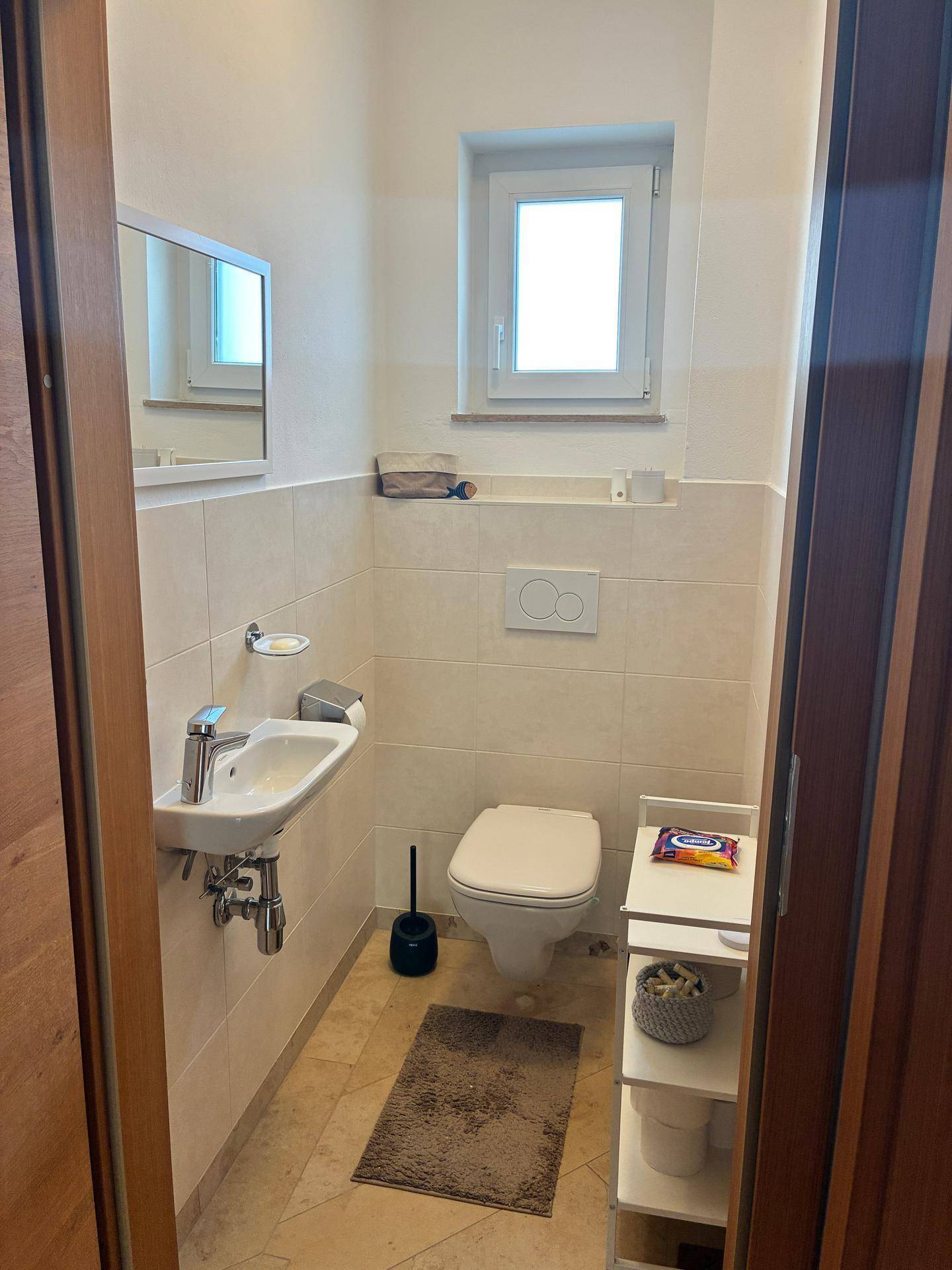 Kleines Gäste-WC mit beigen Fliesen, wandhängendem WC und Fenster für Tageslicht.