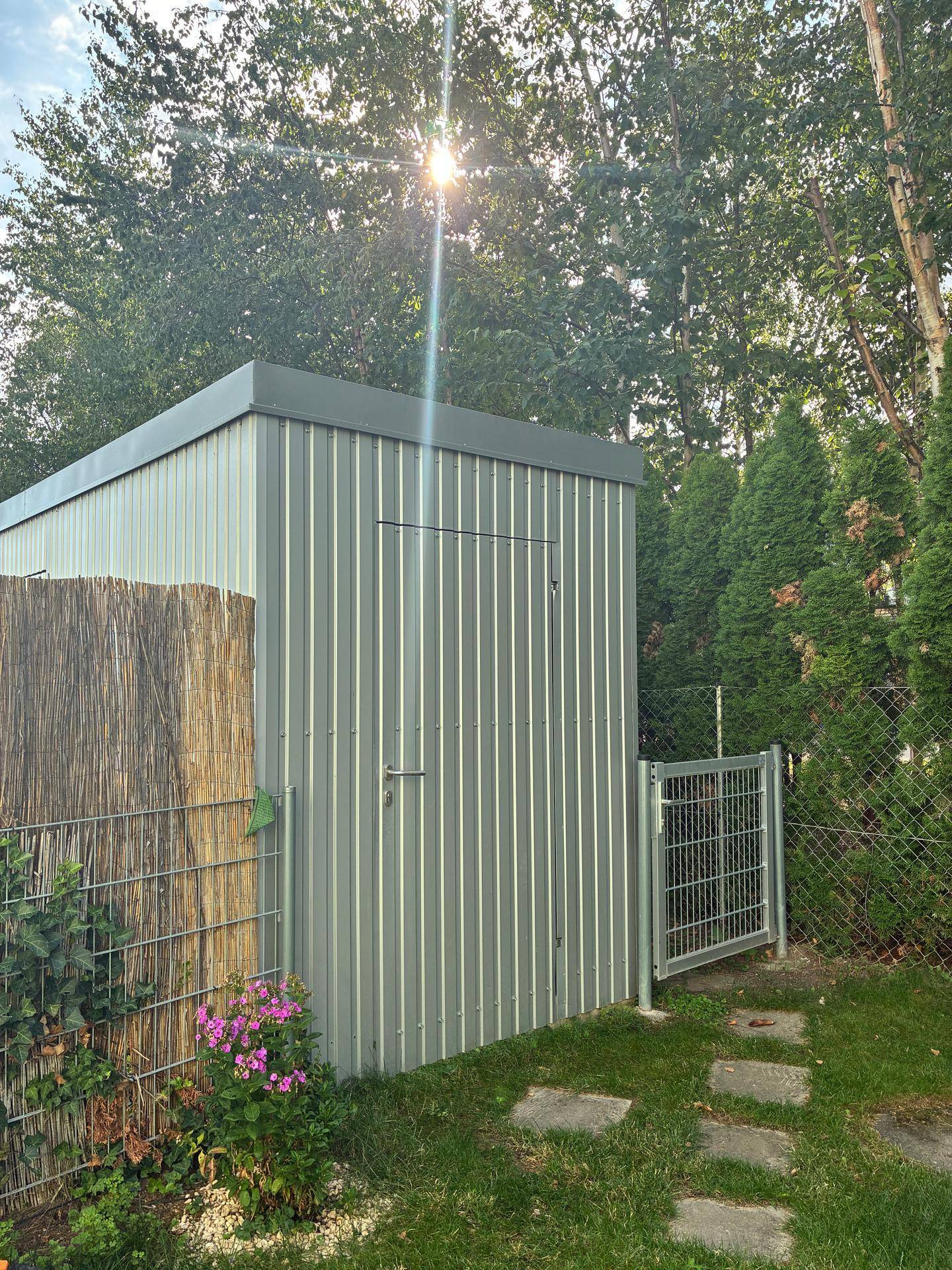 Gartenhaus aus Metall im Grünen, umgeben von Bäumen und einem Zaun.