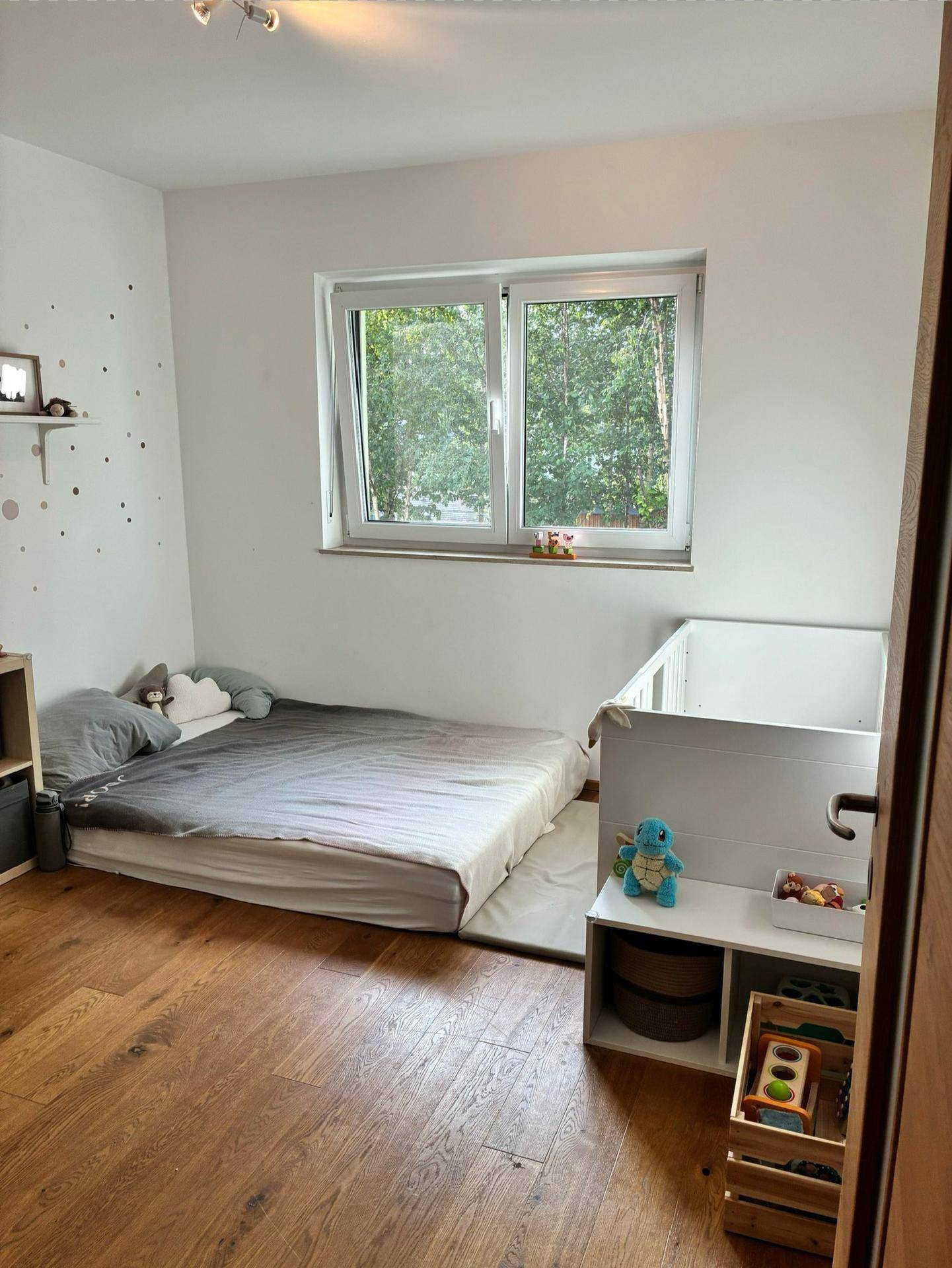 Kinderzimmer mit Holzboden, großem Fenster und Blick auf Bäume.