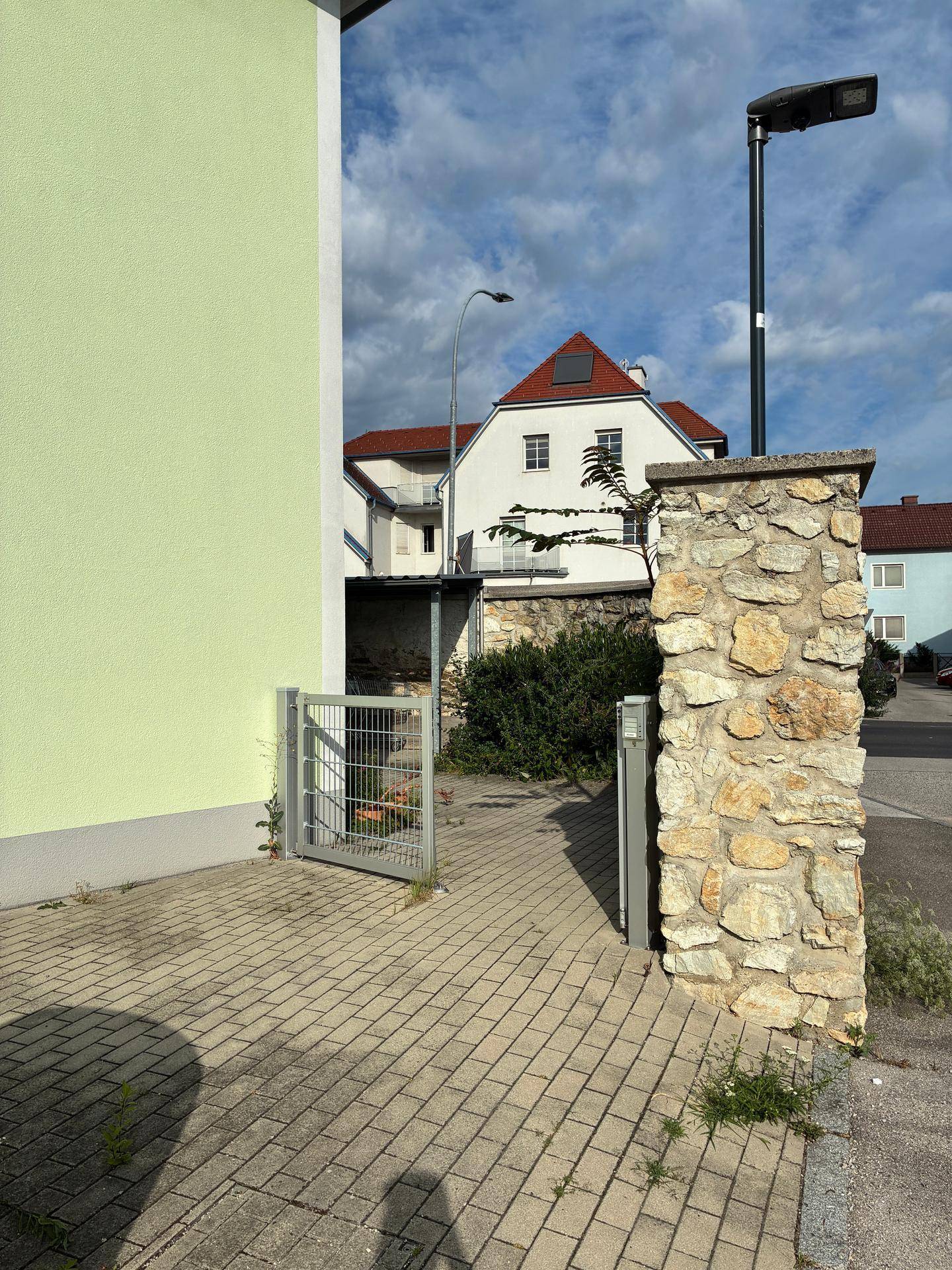 Seitenansicht eines Hauses mit Steinmauer und Blick auf ein weiteres Gebäude im Hintergrund.
