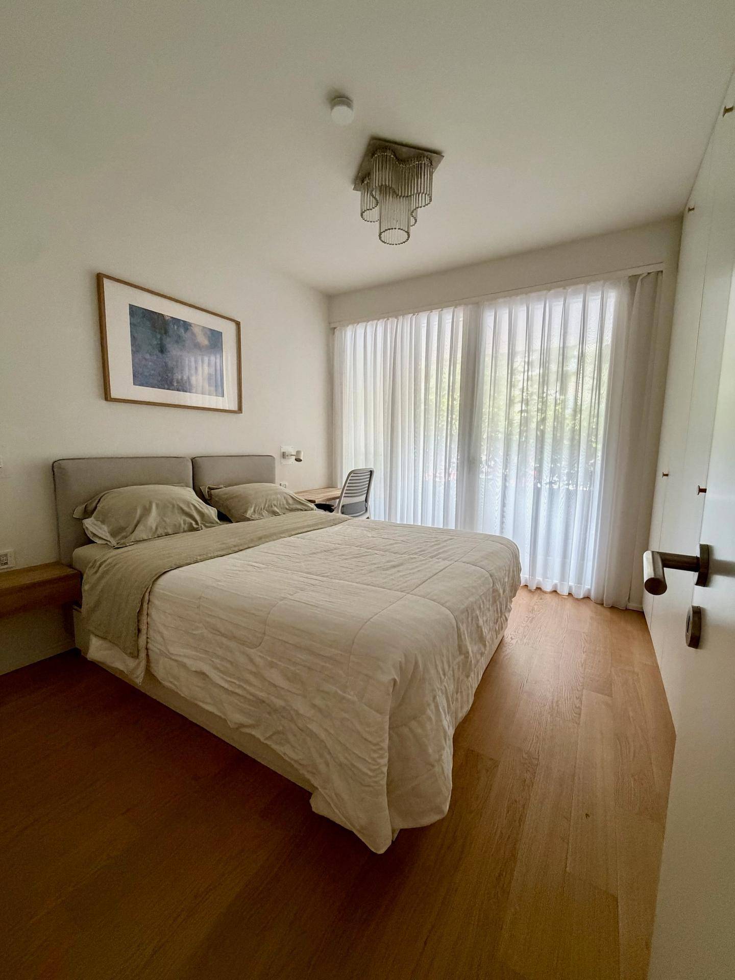 Helles Schlafzimmer mit Doppelbett, Schreibtisch und großen Fenstern mit Vorhängen.