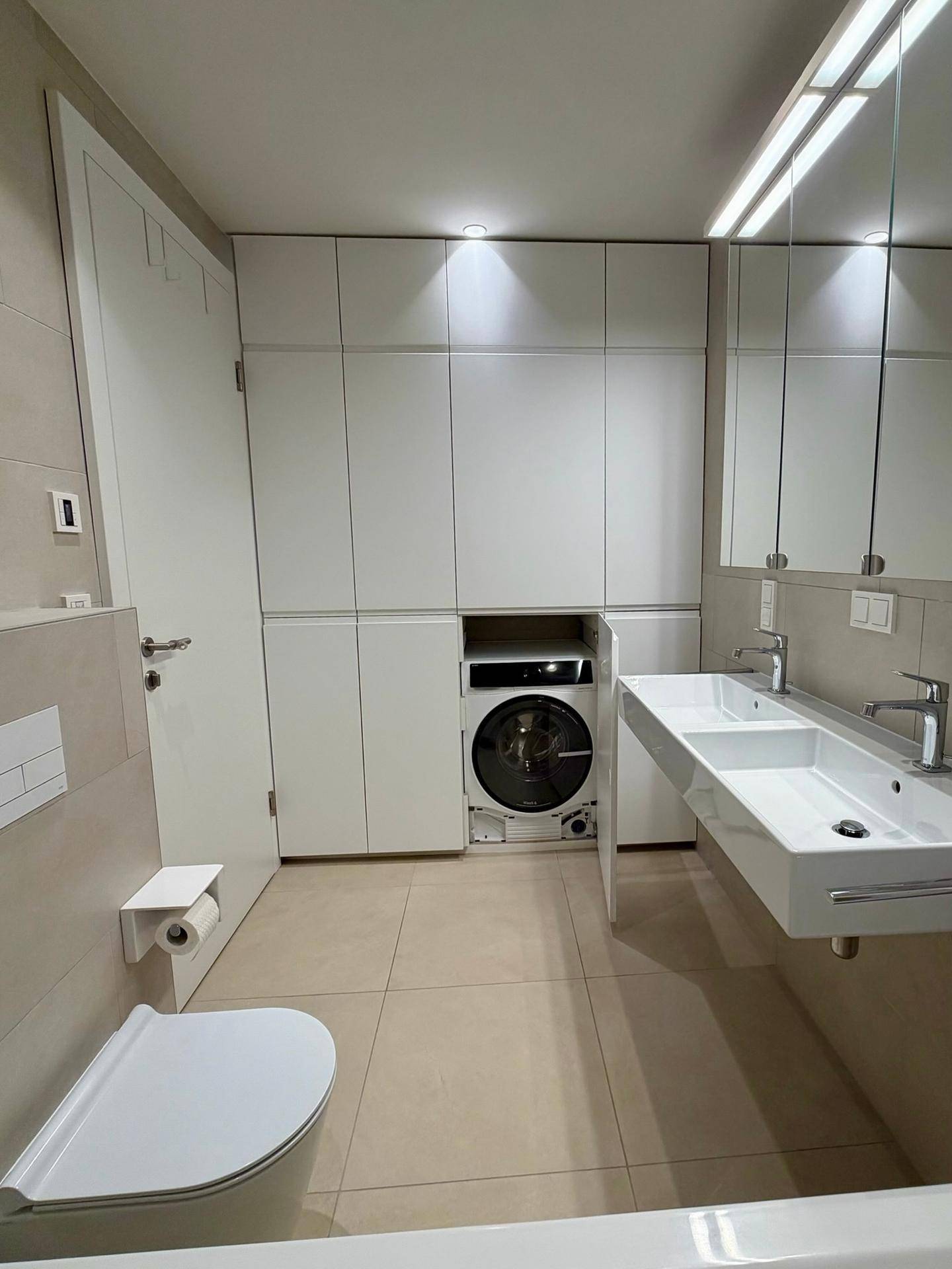 Funktionales Badezimmer mit Doppelwaschbecken, Toilette und integrierter Waschmaschine.