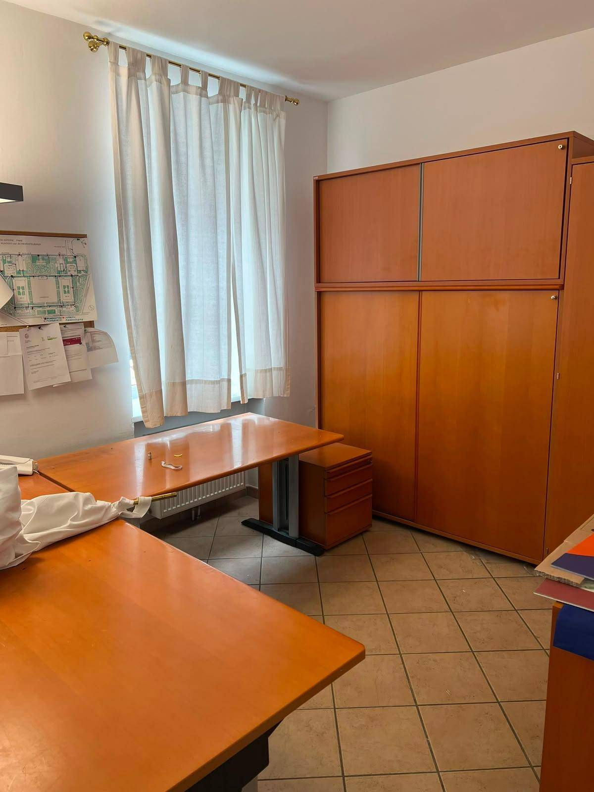 Büro mit zwei Schreibtischen, Holzschränken und einem Fenster mit Vorhängen.