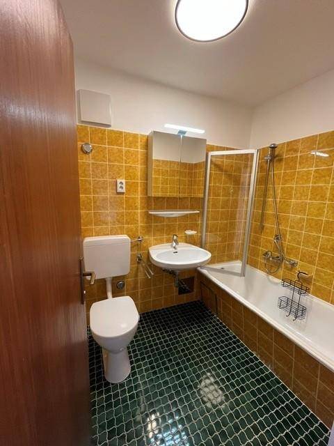 Badezimmer mit gelben Wandfliesen, grünen Bodenfliesen, Badewanne mit Duschabtrennung und WC.