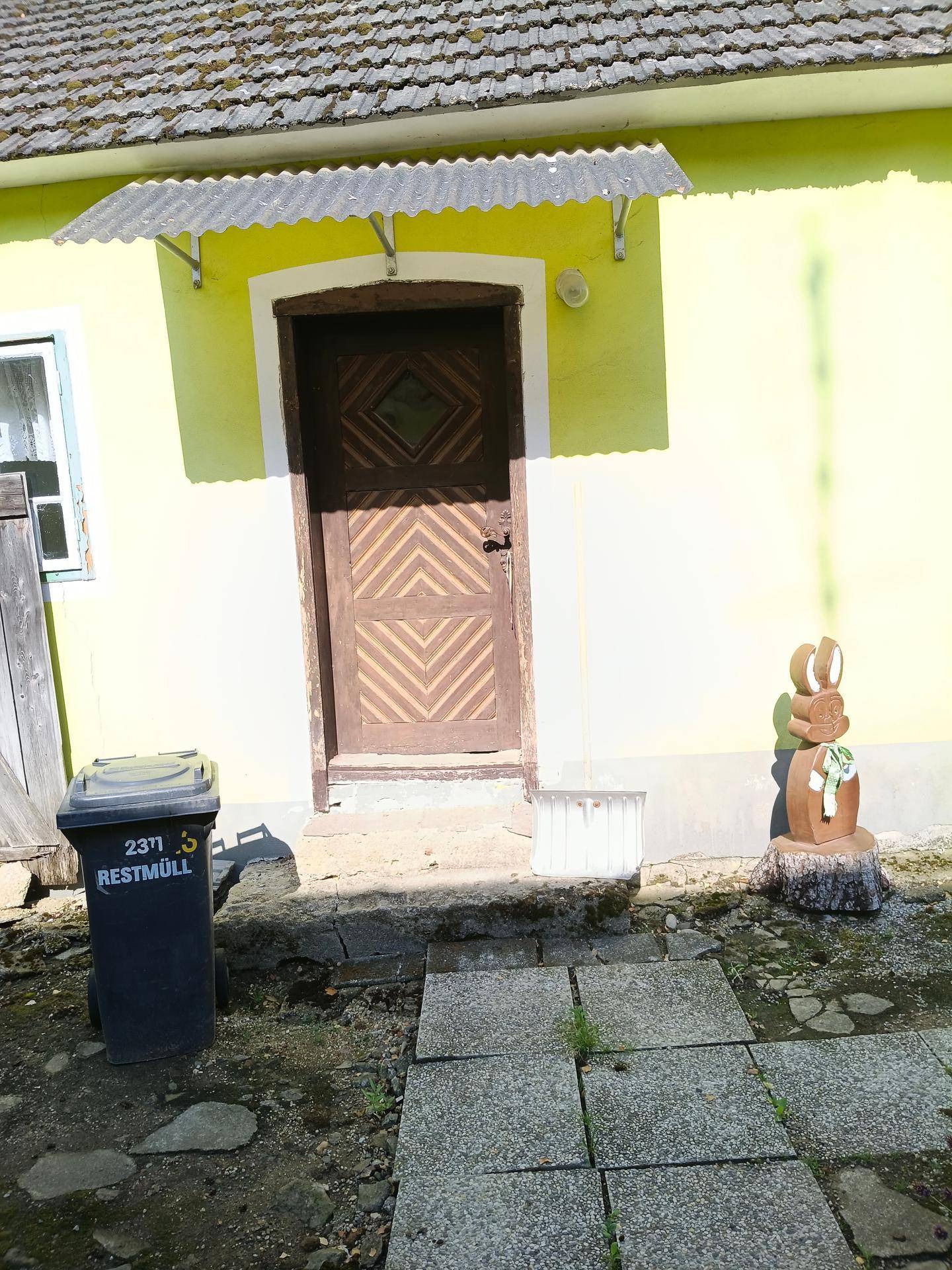 Eingangsbereich eines Hauses mit gelber Fassade, Holztür und Mülleimer.