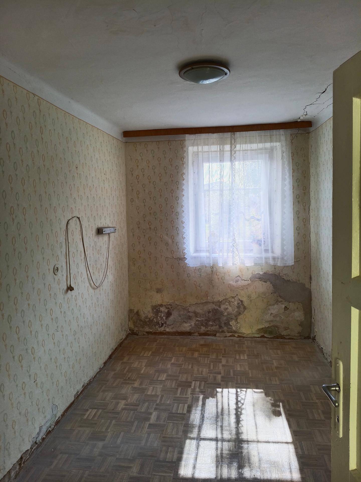 Leeres Zimmer mit abgenutzter Tapete, Parkettboden und einem Fenster.