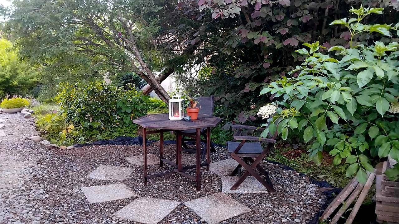 Gemütlicher Gartensitzplatz mit kleinem Holztisch und Stühlen, umgeben von Pflanzen und Kies.