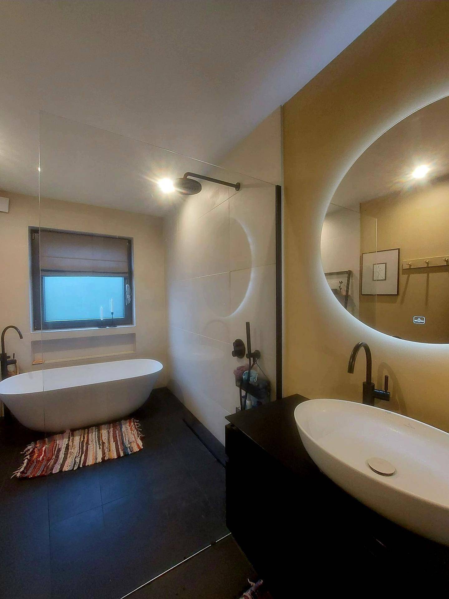 Modernes Badezimmer mit freistehender Badewanne, ovalem Waschbecken und beleuchtetem Spiegel.