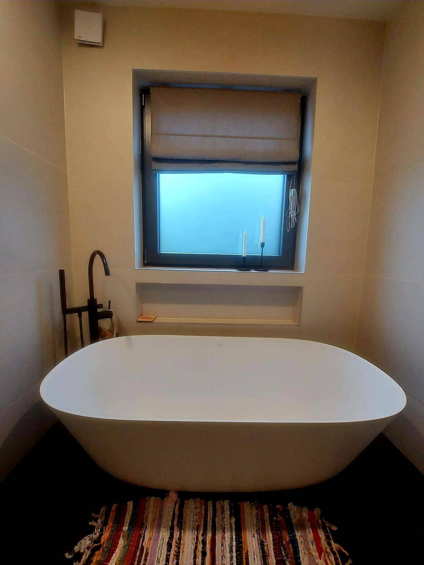 Elegante freistehende Badewanne vor einem Fenster, umgeben von modernen Fliesen.