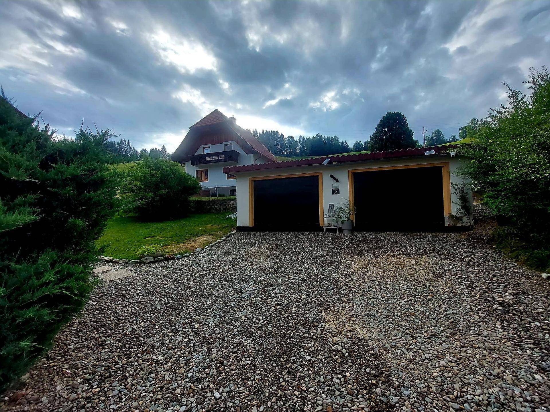 Landhaus mit Doppelgarage und Kiesauffahrt, umgeben von grünen Hügeln unter bewölktem Himmel.
