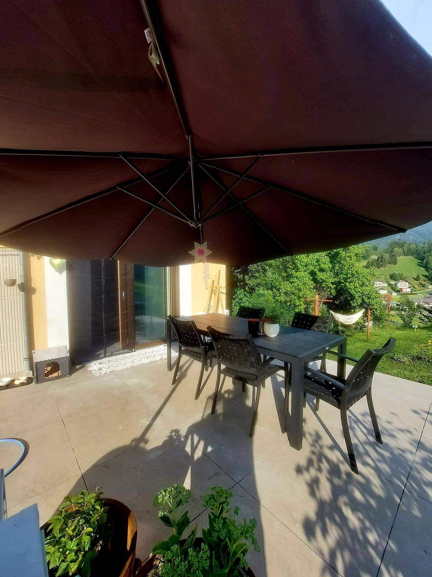Sonnige Terrasse mit Esstisch und Stühlen unter einem großen Sonnenschirm, mit Bergblick.
