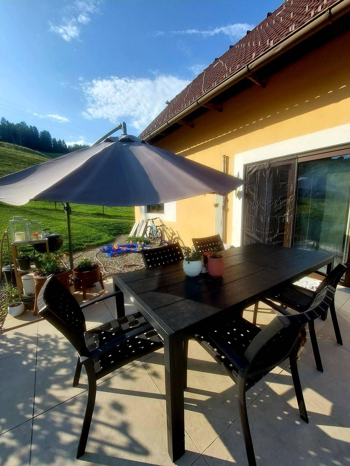 Helle Terrasse mit Esstisch und Sonnenschirm, umgeben von grünen Hügeln und Natur.