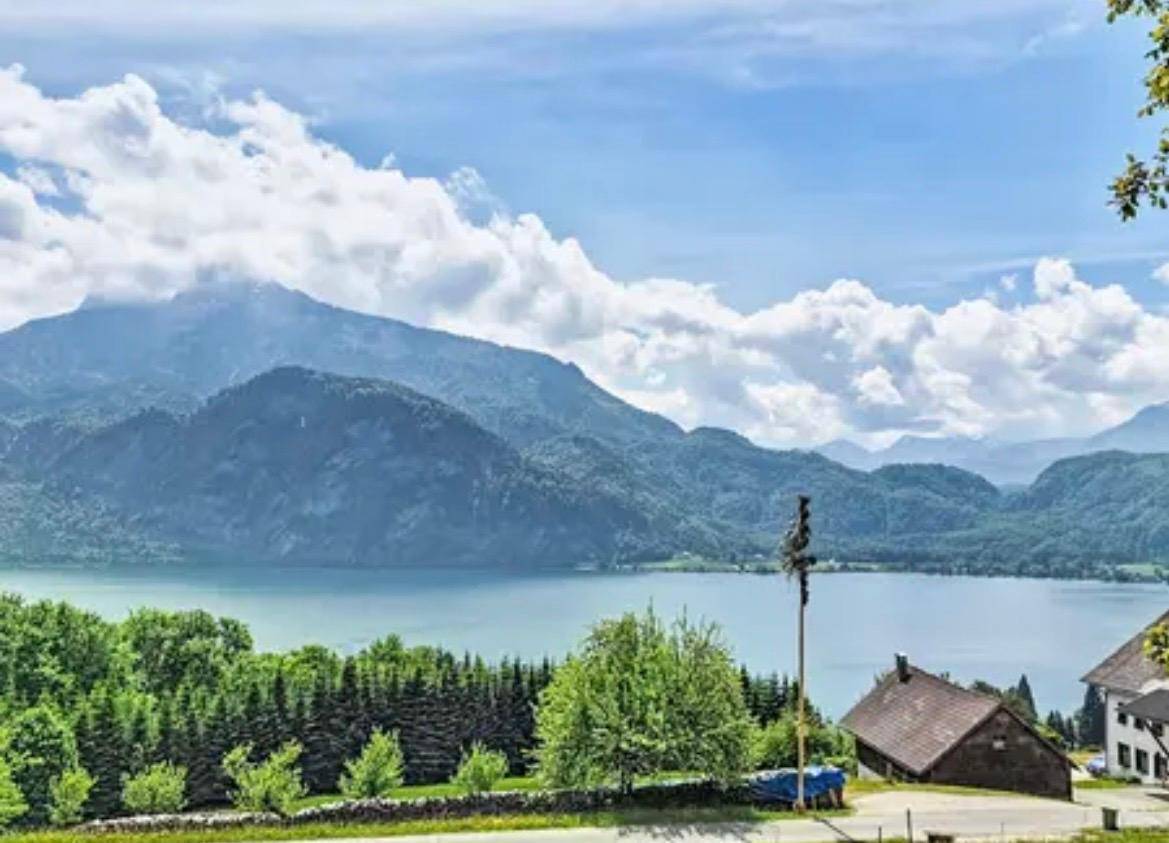 Malerische Landschaft mit See, Bergen und bewaldeten Hügeln unter blauem Himmel.