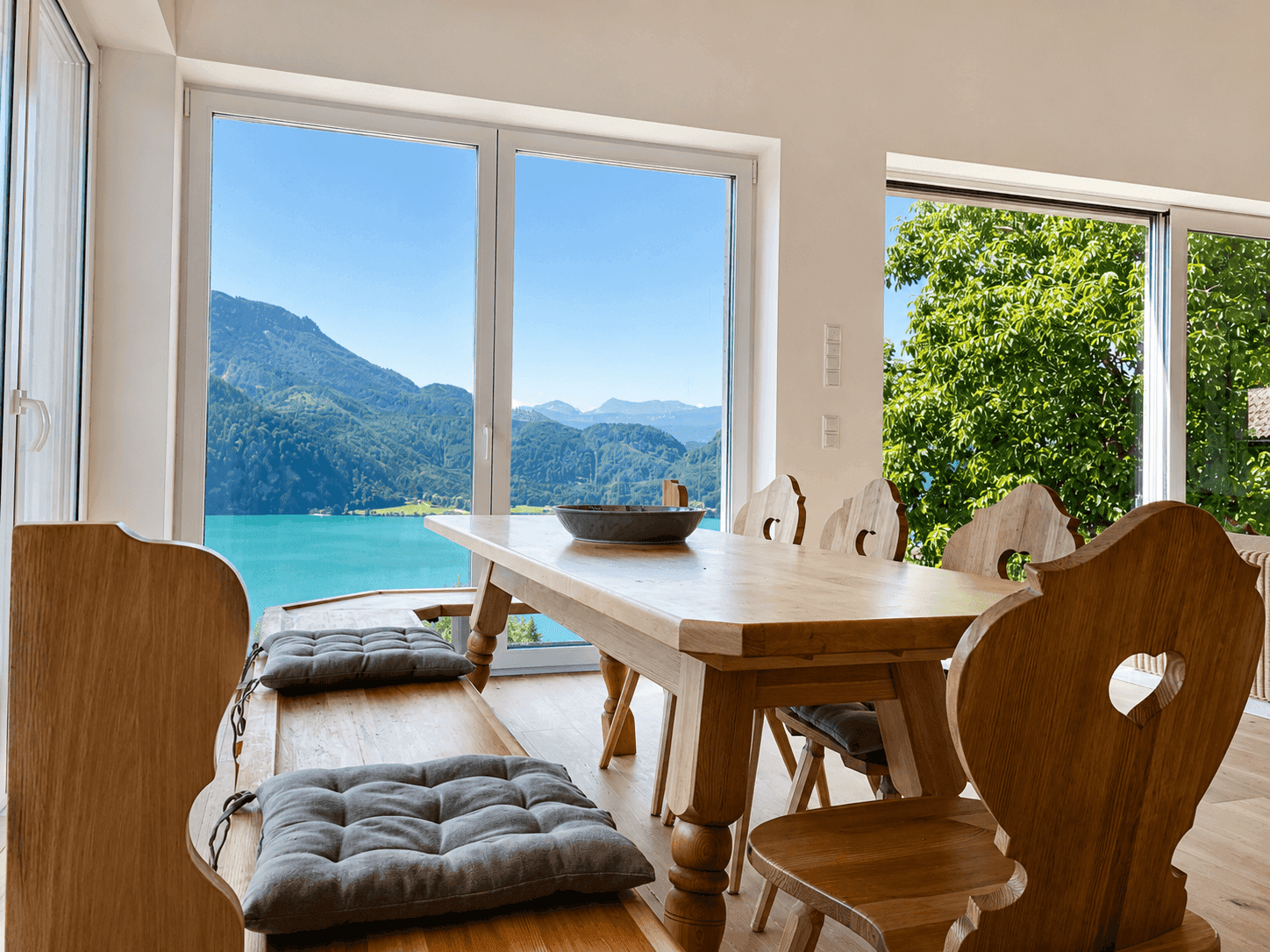 Essbereich mit rustikalem Holztisch und Bänken, umgeben von Fenstern mit Seeblick.