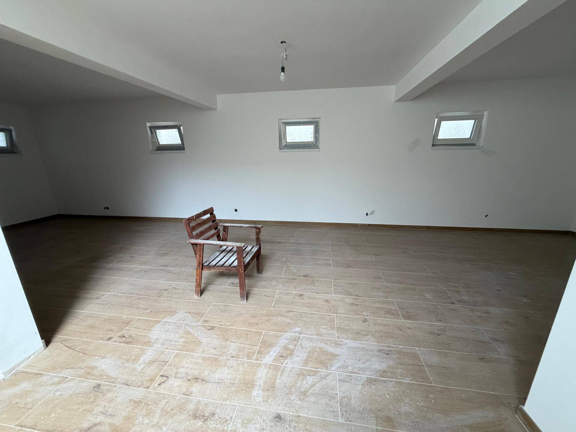 Großer, offener Wohnbereich im Rohbau mit Fliesen in Holzoptik und mehreren kleinen Fenstern.