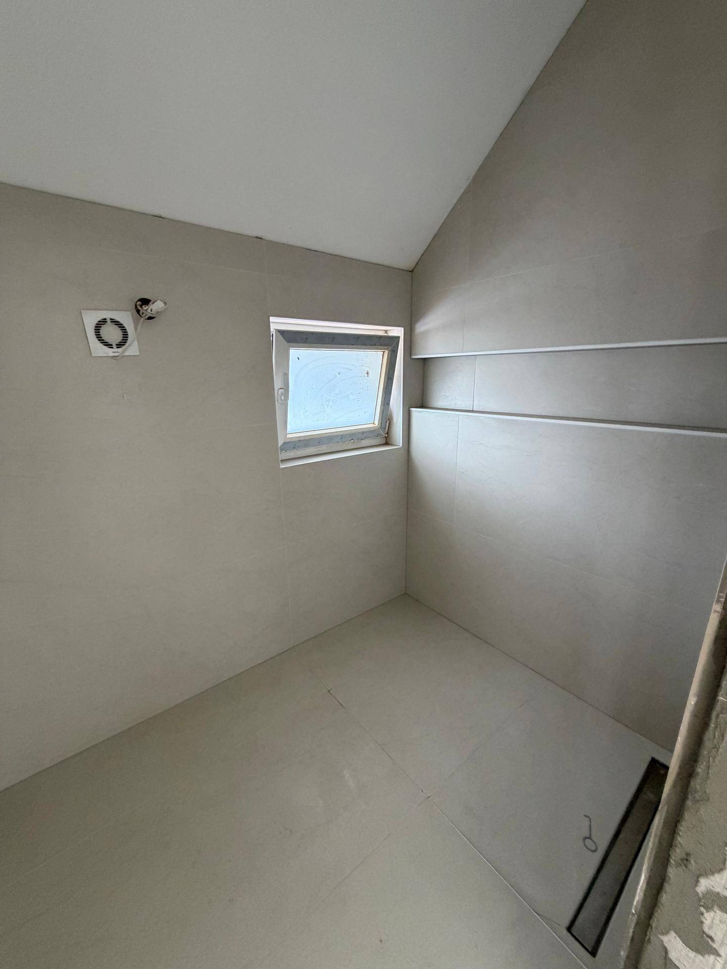 Unfertiges Badezimmer mit hellgrauen Fliesen, einer Nische in der Wand und einem kleinen Fenster.