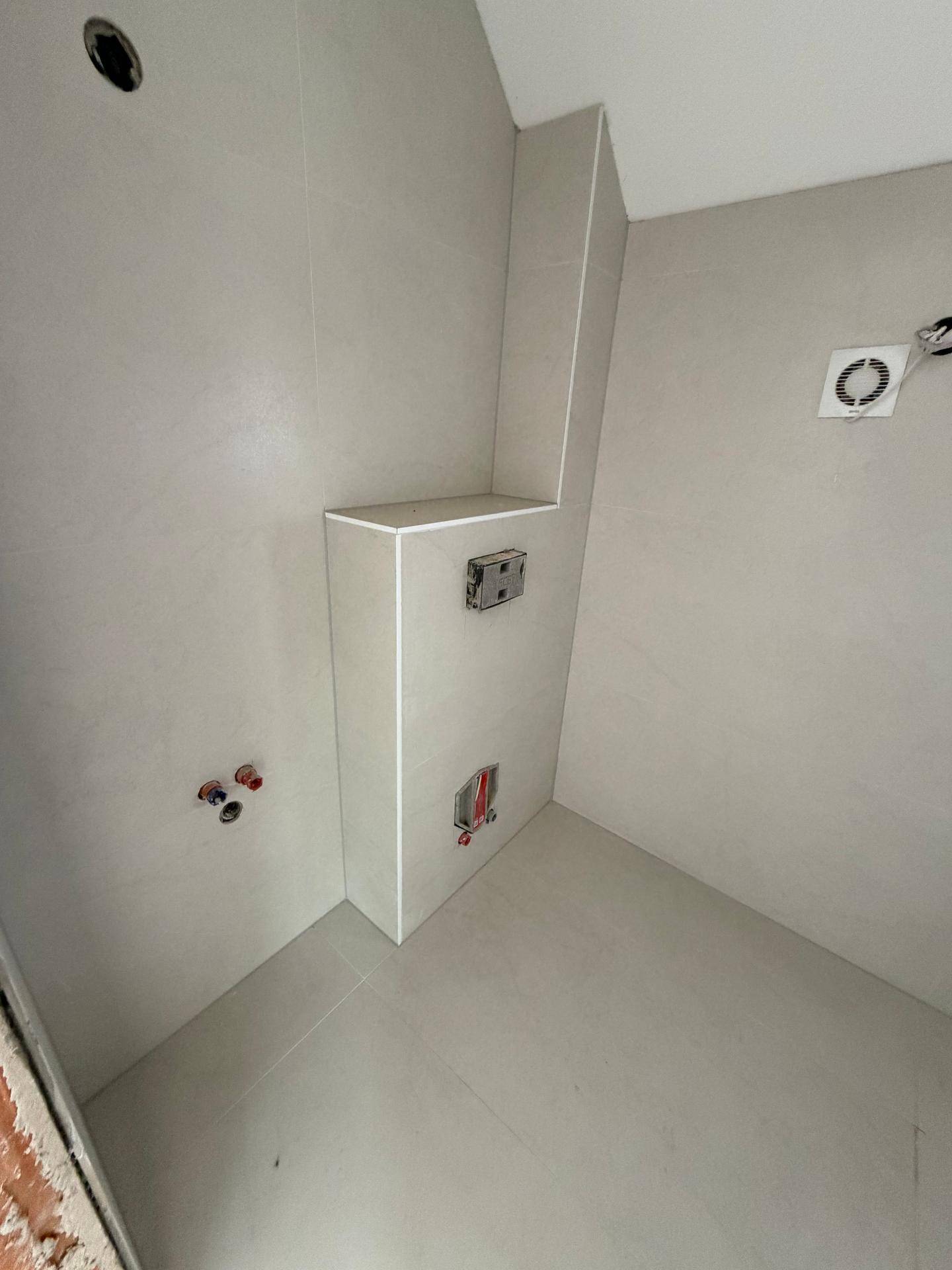 Unfertiges Badezimmer mit hellgrauen Fliesen und sichtbaren Anschlüssen für Sanitärinstallationen.