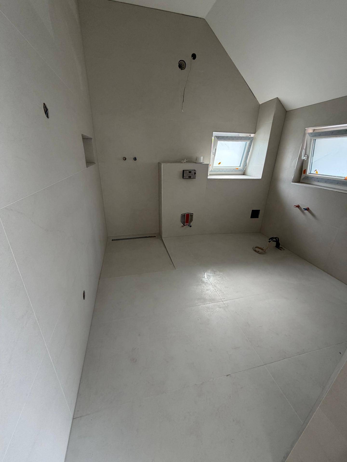 Geräumiges Badezimmer im Rohbau mit hellgrauen Fliesen und mehreren Dachfenstern für natürliches Licht.
