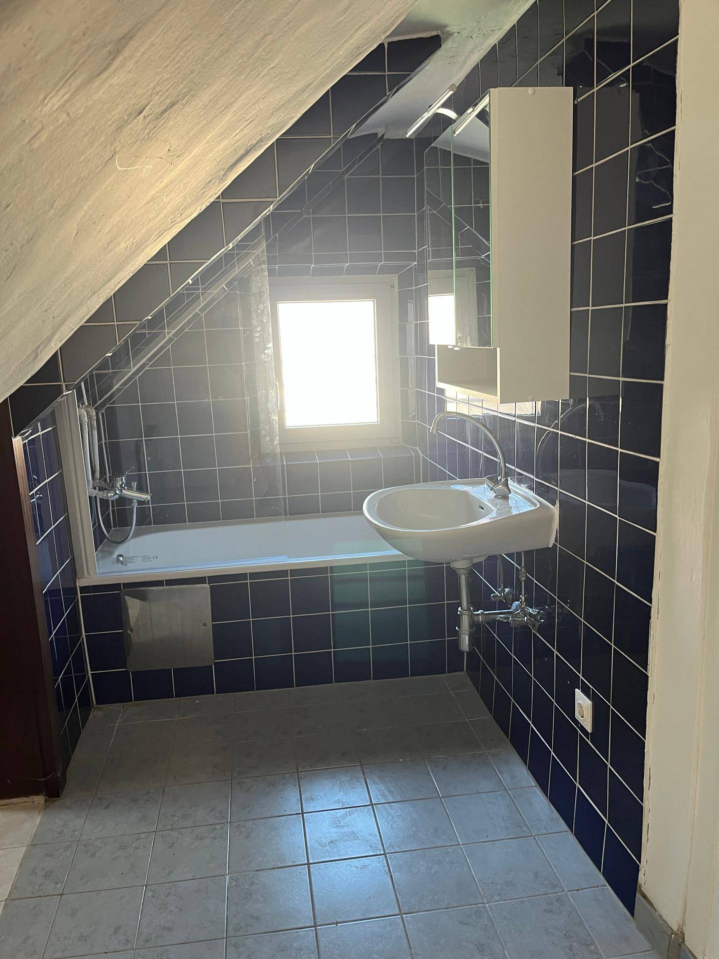 Badezimmer im Dachgeschoss mit dunkelblauen Fliesen, Badewanne, Waschbecken und einem Fenster für Tageslicht.