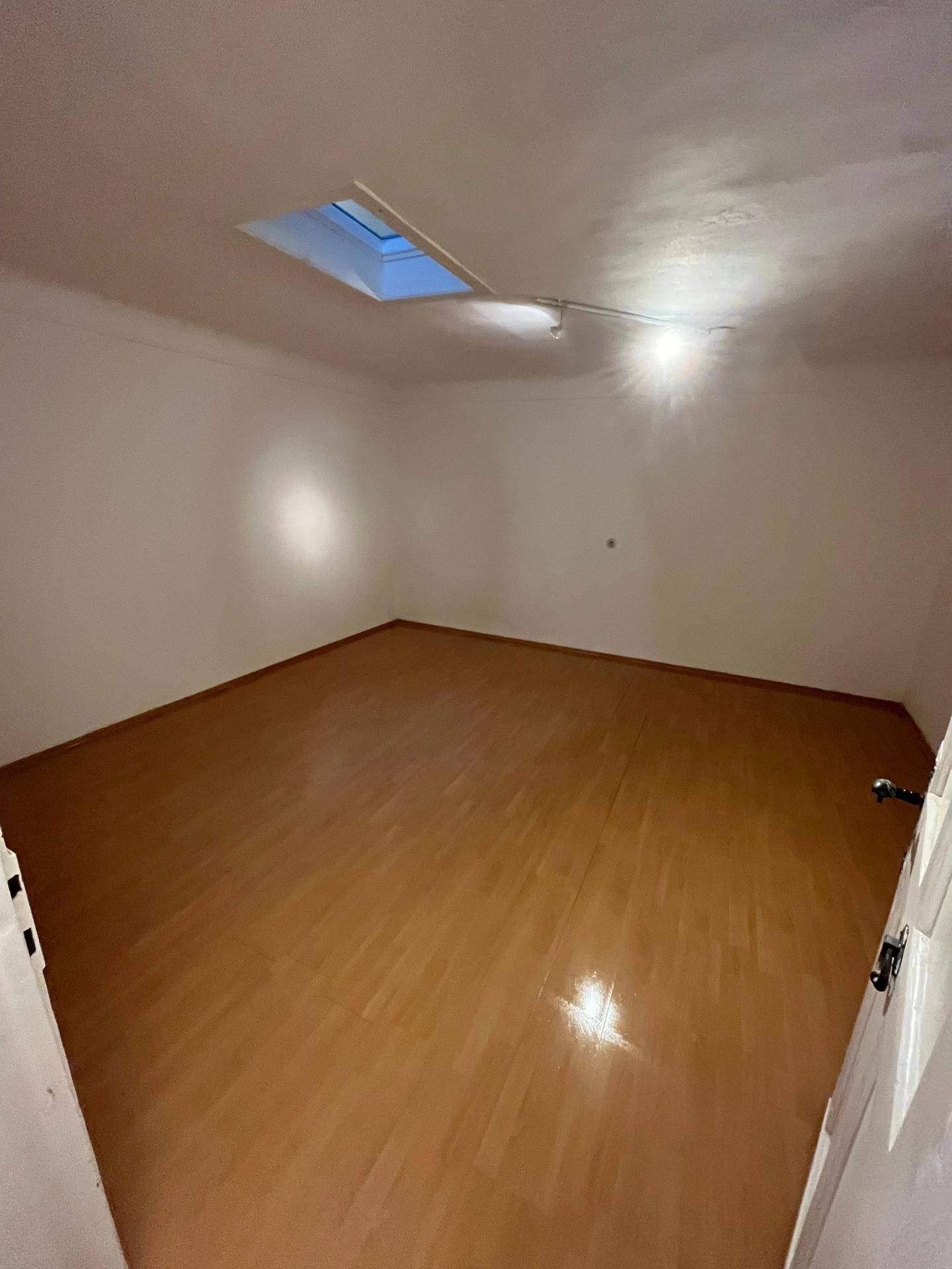 Leeres Zimmer mit Laminatboden, weißen Wänden und einem Dachfenster, das den Raum hell erscheinen lässt.