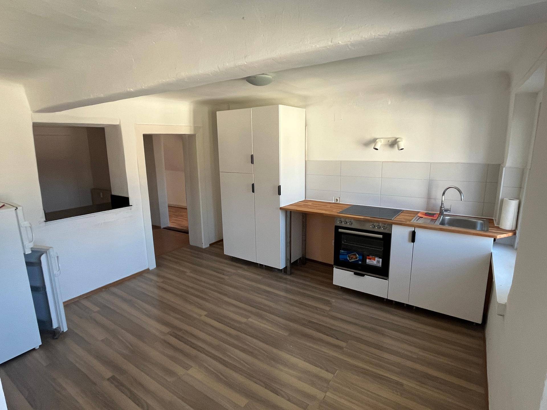 Einfache Küche mit weißen Schränken, Holz-Arbeitsplatte und grundlegenden Geräten, offen zu einem weiteren Raum.