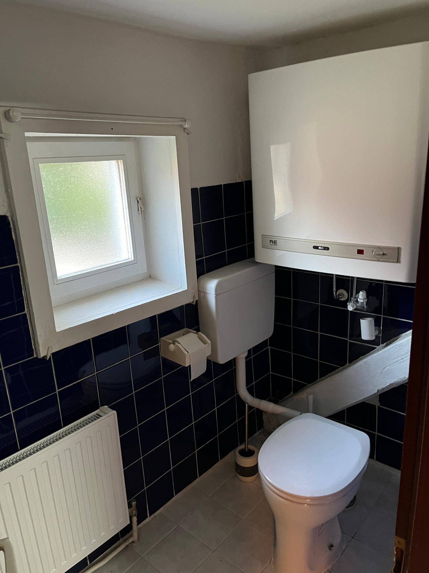 Kleines WC mit dunkelblauen Fliesen, Toilette, Fenster und einem Heizkörper, sowie einem Wandkessel.