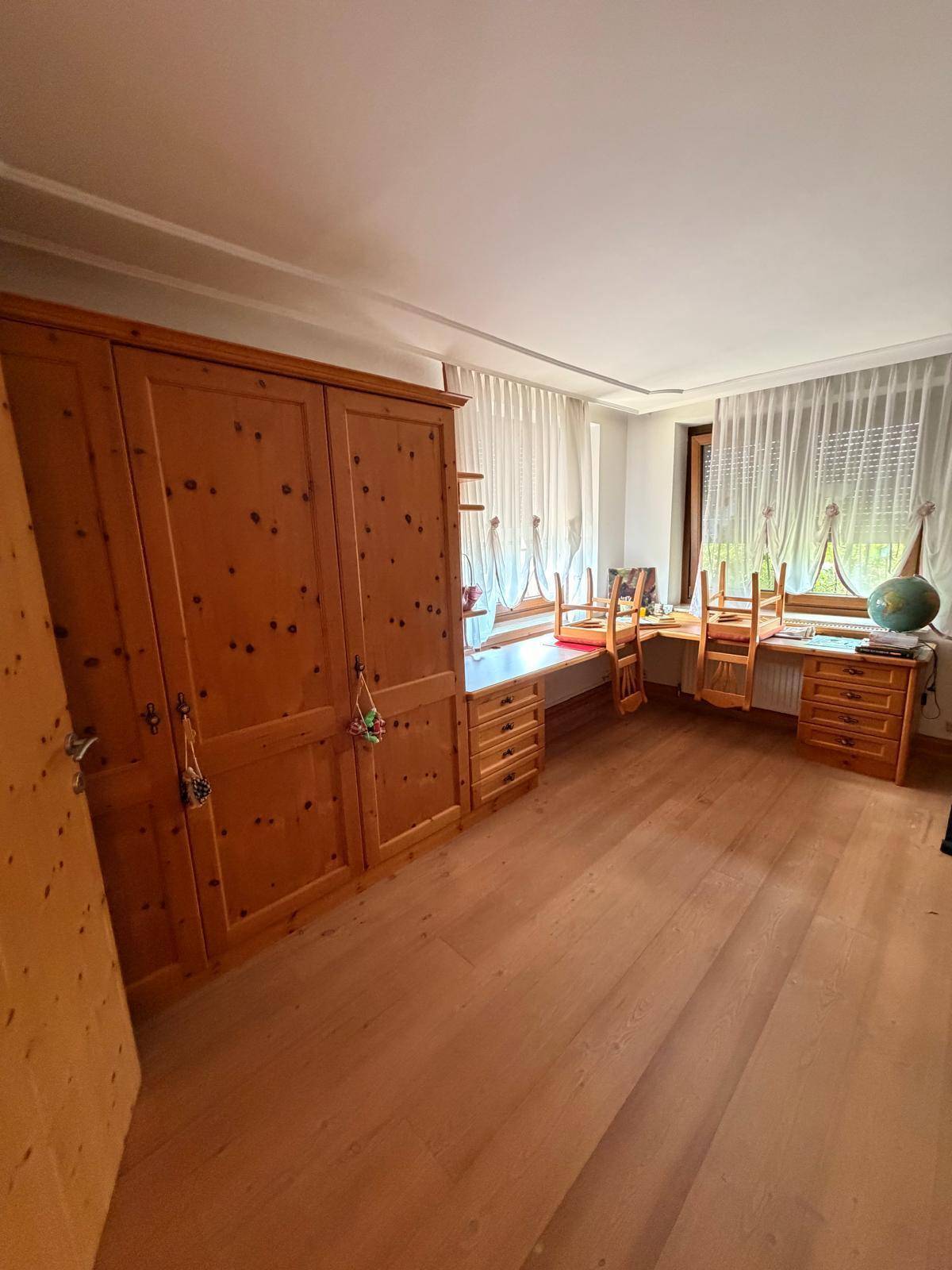 Geräumiges Zimmer mit Holzboden, großem Schrank und Arbeitsbereich am Fenster.