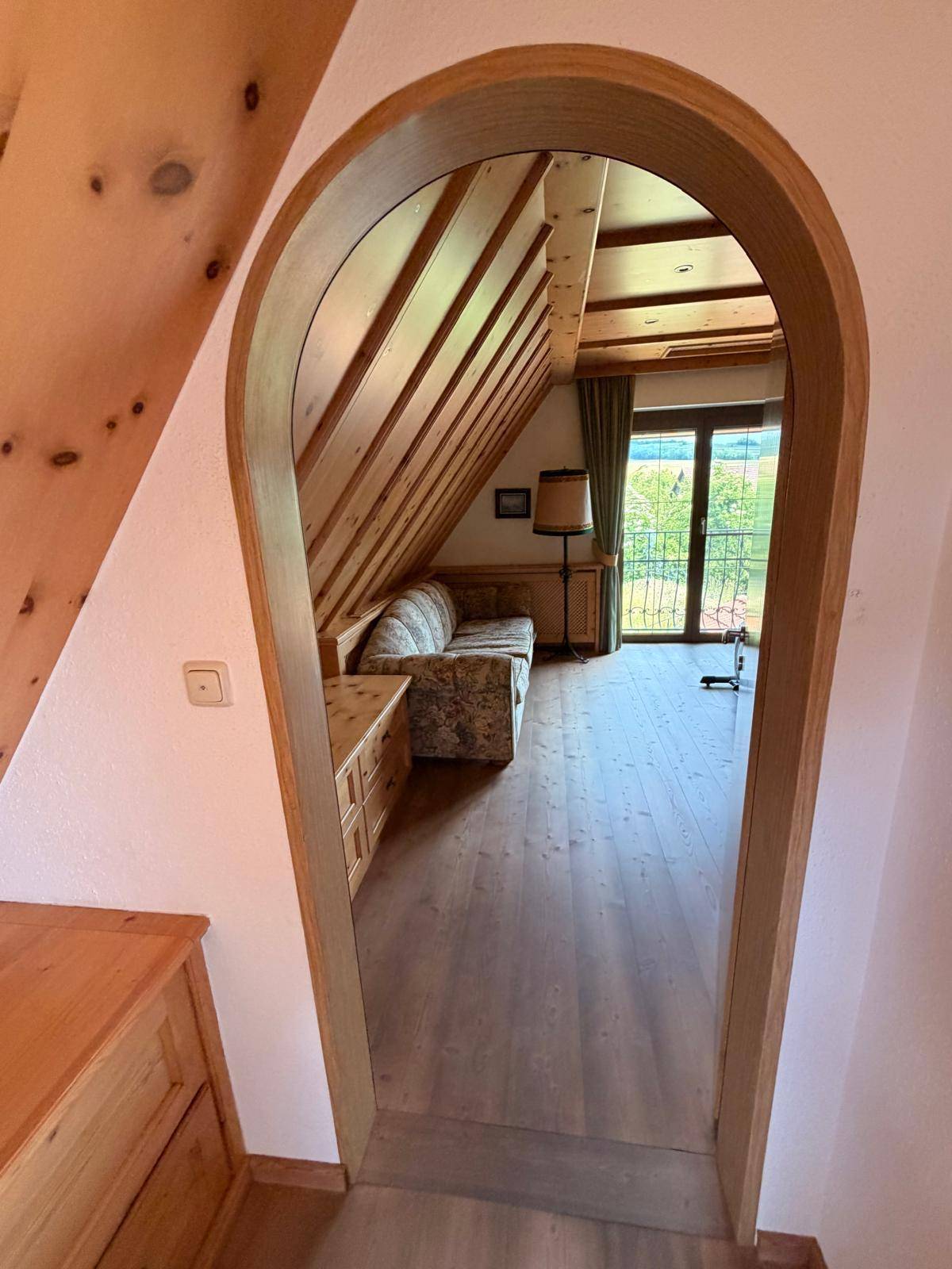 Gemütlicher Wohnbereich im Dachgeschoss mit Holzbalken, Bogenöffnung und Blick ins Grüne.
