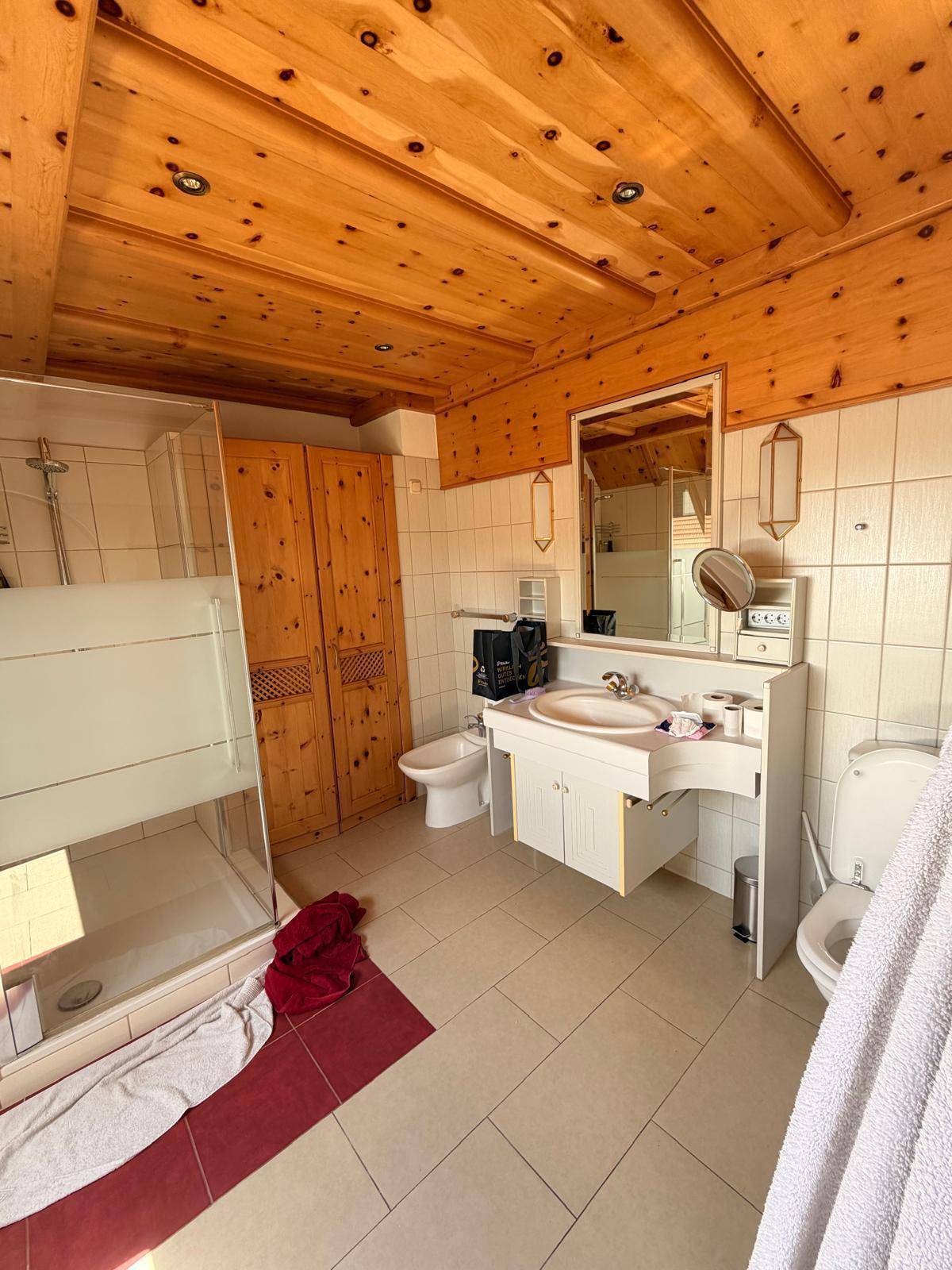 Geräumiges Badezimmer mit Duschkabine, Fliesenboden und Holzschränken unter der Holzdecke.