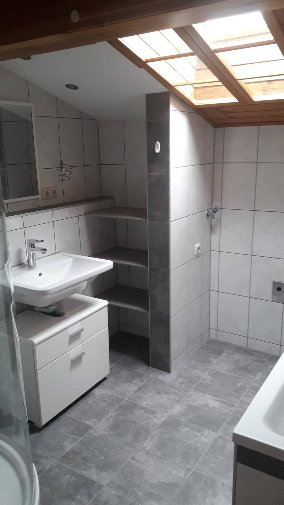Helles Badezimmer mit grauen Fliesen, Waschbecken, Dusche und einem Dachfenster für natürliches Licht.
